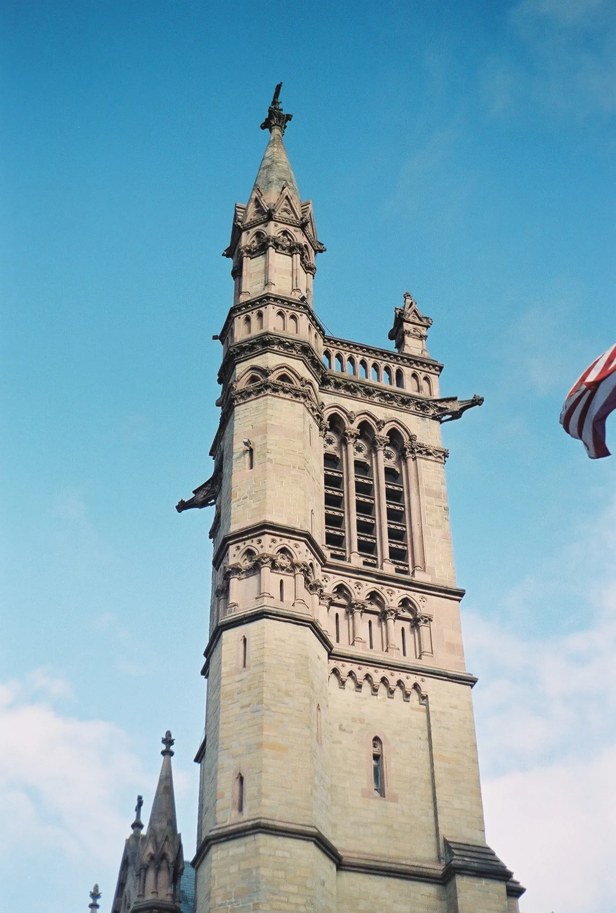 A tall stone tower with ornate arches and a figure at the top, flag visible at right edge, shot from below against clear blue sky.